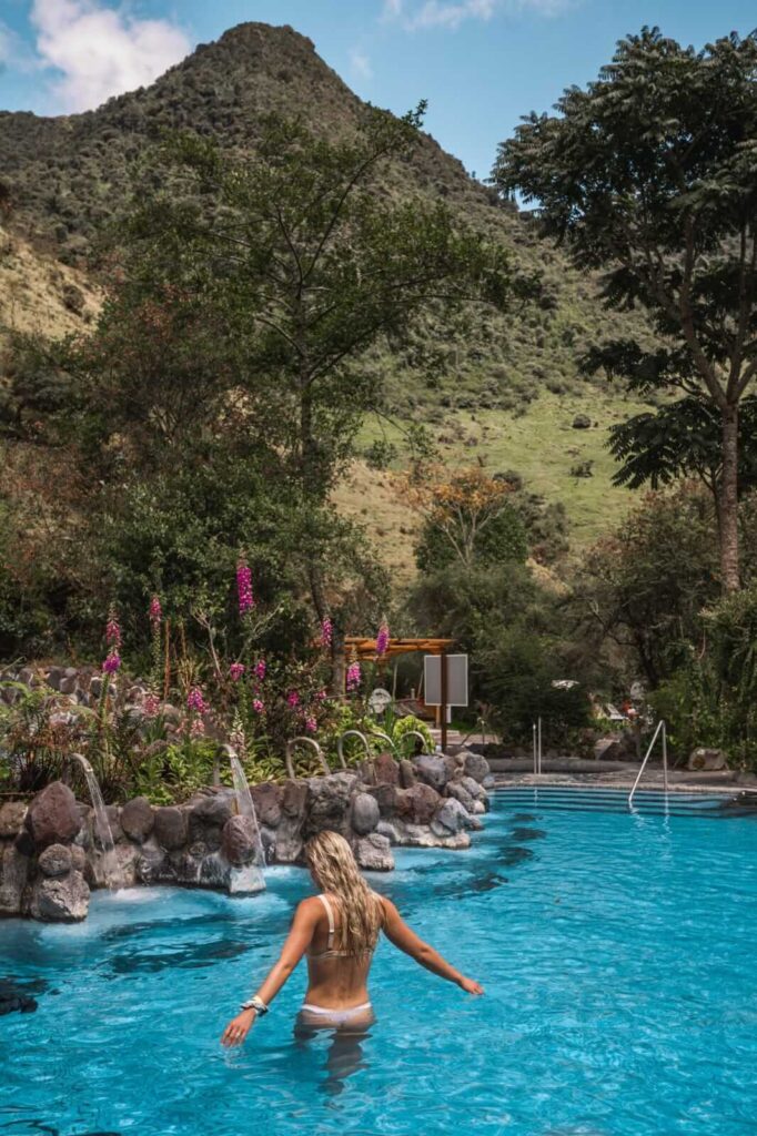 Woman standing in a blue thermal pool with small waterfalls and foxgloves at Termas de Papallacta